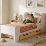 Letto per bambini FLOU D con cassetto, bianco e legno naturale con mensola per libri, una bambina legge un libro sul letto.