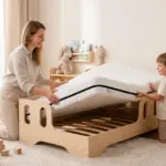 Letto per bambini TILA 1 in legno naturale con materasso, mamma e figlio sistemano il materasso sulla rete a doghe in una cameretta luminosa.