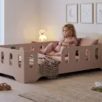 Letto per bambini TILA 2 color cachemire con sponde, bambina seduta che abbraccia un cuscino in una cameretta calma e accogliente.