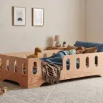 Letto per bambini TILA 2 in legno naturale con sponde di sicurezza, bambina che dorme avvolta in una coperta blu nella sua cameretta.