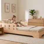 Letto per bambini TILA 6 in legno naturale con barriera traforata, bambina che legge un libro in una cameretta luminosa.