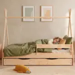 Letto a capanna TIPI D in legno naturale con cassetto e sponda, bambina che dorme sotto un piumone verde oliva.