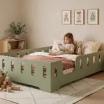 Letto per bambini TILA 2 verde con barriere, bambina che legge un libro tra i cuscini in una cameretta luminosa.