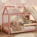 Letto a capanna MILO rosa con baldacchino e barriera in legno naturale, bambina che legge un libro tra i cuscini.