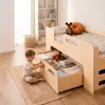 Letto PIPI in legno naturale con cassetto estraibile aperto, bambino che sistema giochi e libri nello scomparto.