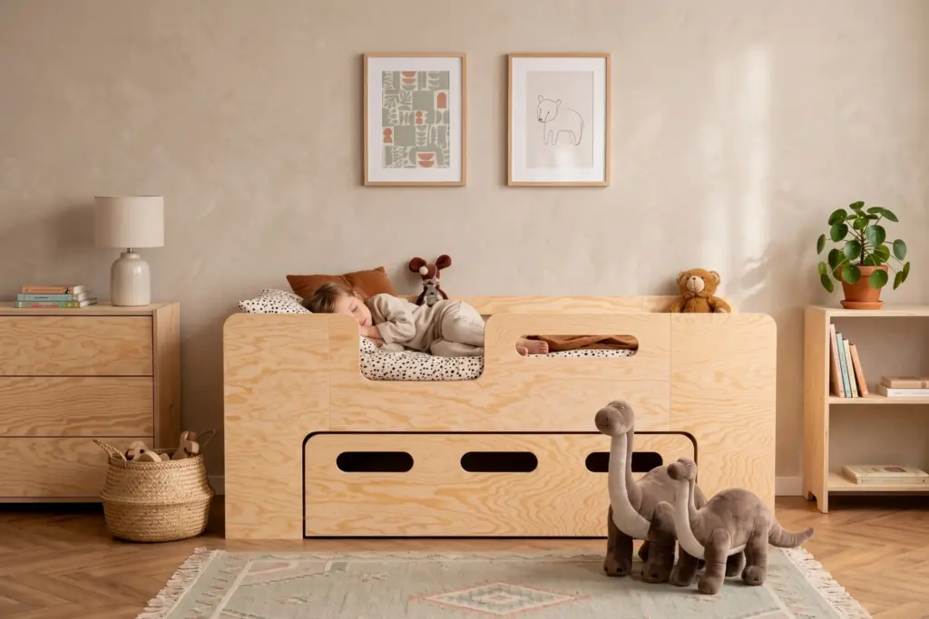 Letto per bambini PIPI in legno naturale con cassetto e ingresso laterale, bambina che dorme in una cameretta luminosa.