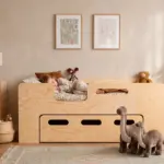Letto per bambini PIPI in legno naturale con cassetto e ingresso laterale, bambina che dorme in una cameretta luminosa.