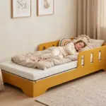 Letto per bambini TILA 6 giallo senape con barriera, bambina che dorme sul materasso in una cameretta luminosa.