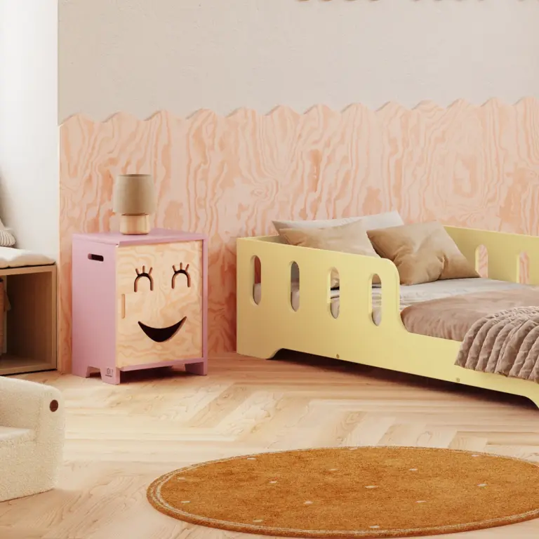 Comodino Kapi rosa con frontale decorativo sorridente, accanto al letto per bambini Tila 6 in tonalità burro.