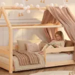 Letto a capanna MILO in legno naturale di pino con sponda e baldacchino, bambina che legge un libro tra i cuscini.