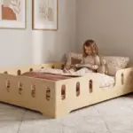 Lettino TILA 2 in legno naturale con sponde ed entrata laterale, bambina che legge un libro appoggiata ai cuscini